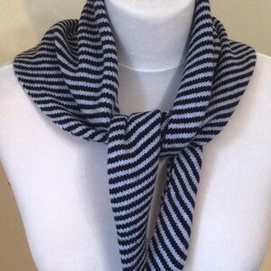 May include: A navy and light grey striped scarf, knotted at the front. The scarf is displayed on a white mannequin. The knitted scarf has a repeating pattern of horizontal stripes. The scarf is a neck accessory.