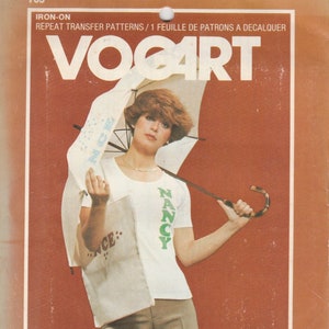 May include: A woman wearing a white shirt with a green "NANCY" graphic and brown pants holds an umbrella. The image is from a vintage iron-on transfer pattern sheet. The text "VOOART" is at the top of the image. The text "Full Block Alphabet Included" is at the bottom of the image.