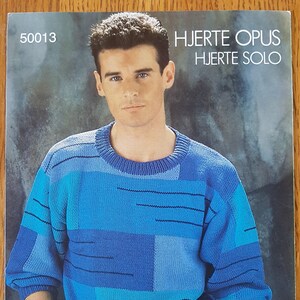 May include: A blue and turquoise patterned sweater with a crew neck. The sweater is made of a soft, textured yarn. The model is wearing blue jeans and has a casual, relaxed look. The text "HJERTETOPUS HJERTESOLO" is printed in white on a black background at the top of the image. The text "HJERTEGARN danish design" is printed in white on a red heart at the bottom of the image.