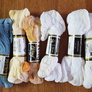Op de afbeelding: Assortiment van Daisy merk gemerceriseerd haakgaren in verschillende kleuren, waaronder groen, blauw, crème en wit. De spoelen zijn gelabeld met de merknaam, het garentype en de kleur.
