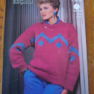 May include: A pink sweater with a blue geometric pattern. The sweater is knitted and has a crew neck. The model is wearing blue jeans and has short brown hair.