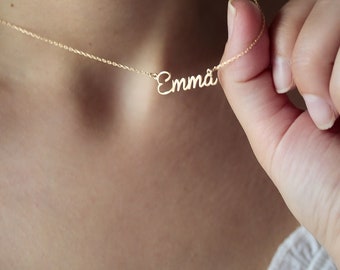 May include: A delicate gold-colored necklace with the name "Emma" in cursive script. The necklace is held up against a person's neck, showcasing the pendant's design and the chain's fine details. The necklace is a personalized jewelry item.