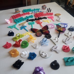 May include: A collection of colorful cake toppers and phone ring holders. The toppers include butterfly, flower, and text designs in various colors like red, blue, and yellow. The phone ring holders are in various shapes and colors.