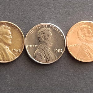 May include: Three U.S. Lincoln pennies are shown against a dark backdrop. The coins are dated 1968 and 1982, with the profile of Abraham Lincoln visible. The coins are in varying states of wear, showcasing their age and history.
