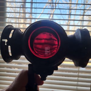 Puede incluir: Linterna vintage negra con lente roja. La linterna tiene un cuerpo redondo con asa y soporte. La lente es de color rojo brillante y la linterna parece ser de metal.