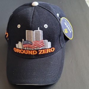 May include: Navy blue baseball cap with embroidered design. The design includes the text "GROUND ZERO" in yellow, a depiction of the Twin Towers, and an American flag. The cap has a white button on top and a tag.
