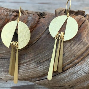May include: A pair of gold-colored earrings. Each earring features a circular top with a triangle cut-out and three dangling, elongated, rectangular pendants. The earrings are displayed on a piece of weathered wood.