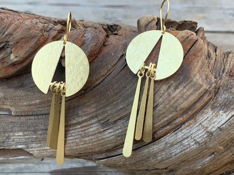 May include: A pair of gold-colored earrings. Each earring features a circular top with a triangle cut-out and three dangling, elongated, rectangular pendants. The earrings are displayed on a piece of weathered wood.