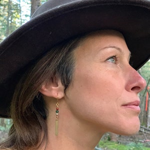 May include: A woman wearing a brown felt hat and a tan sweater with gold earrings featuring a rectangular charm and green, brown, and white beads.