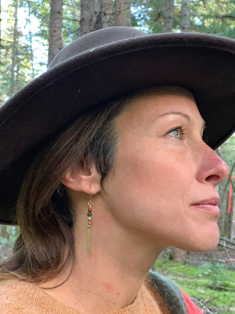May include: A woman wearing a brown felt hat and a tan sweater with gold earrings featuring a rectangular charm and green, brown, and white beads.