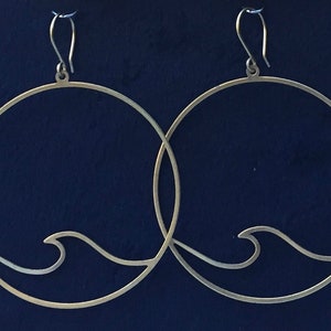 May include: A pair of silver-toned hoop earrings. Each earring features a circular design with a wave detail at the bottom. The earrings have a simple hook closure and are set against a dark blue background.