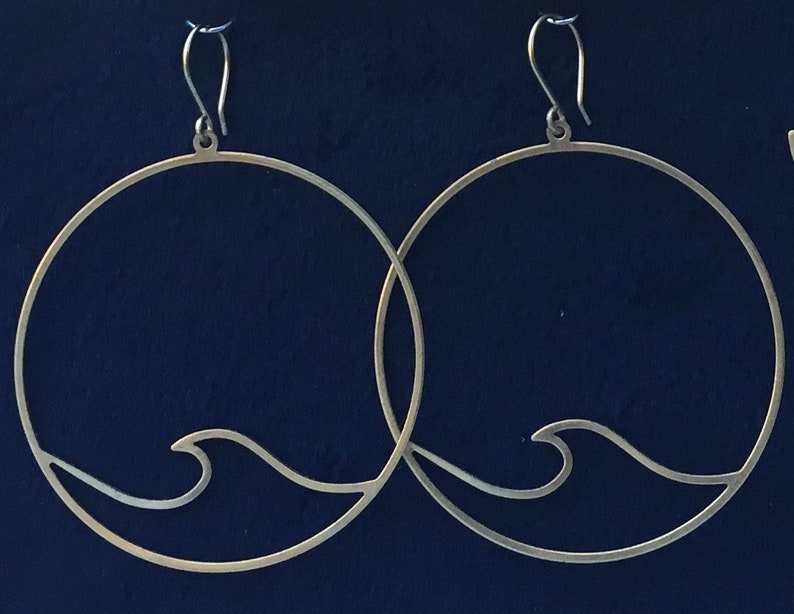 May include: A pair of silver-toned hoop earrings. Each earring features a circular design with a wave detail at the bottom. The earrings have a simple hook closure and are set against a dark blue background.