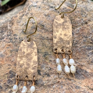 May include: A pair of handcrafted earrings with a textured, rectangular gold-toned metal base. Each earring features three dangling, irregular-shaped white pearls, suspended by gold-colored wire. The earrings have a hook closure.