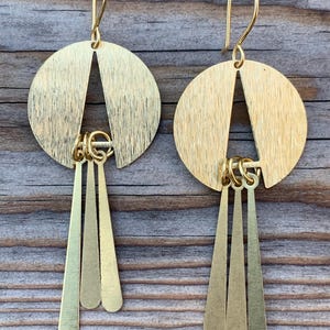 May include: A pair of gold-colored earrings. Each earring features a circular top with a triangular cutout and three dangling, elongated teardrop shapes. The earrings have a hook closure and are displayed against a wooden background.