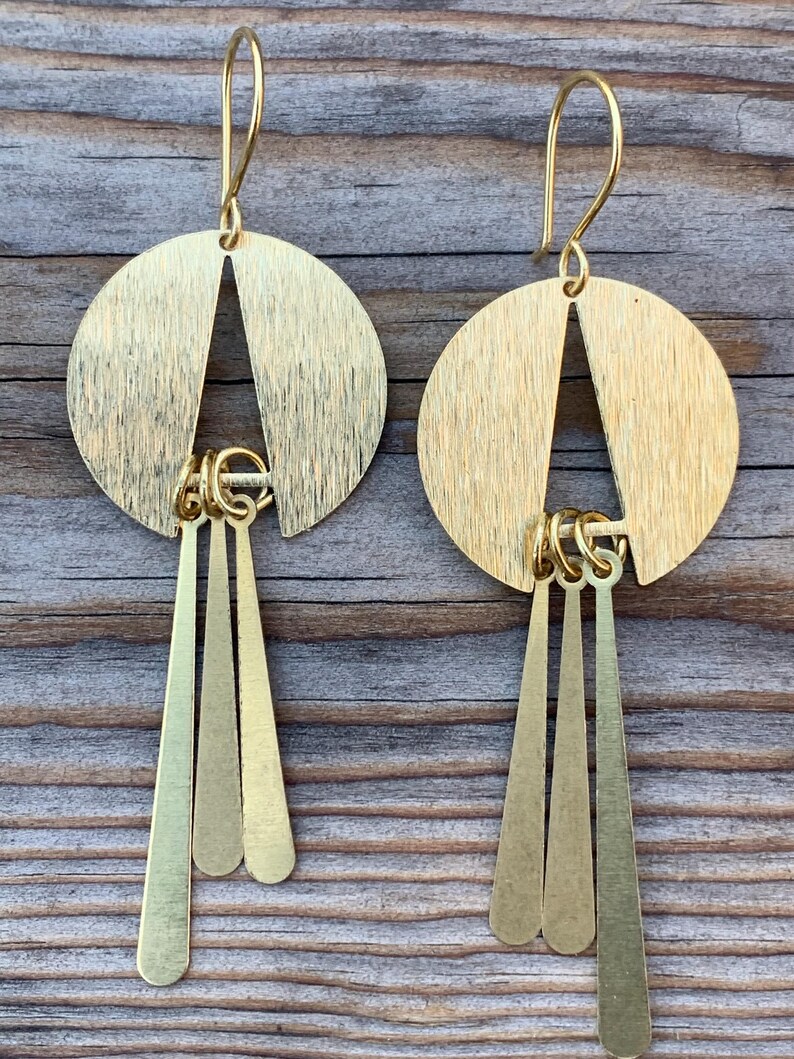 May include: A pair of gold-colored earrings. Each earring features a circular top with a triangular cutout and three dangling, elongated teardrop shapes. The earrings have a hook closure and are displayed against a wooden background.