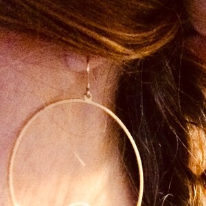 May include: Gold-colored hoop earrings with a wave design. The earrings are circular with a delicate wave detail cut out of the metal. The earrings are shown against a background of brown hair.