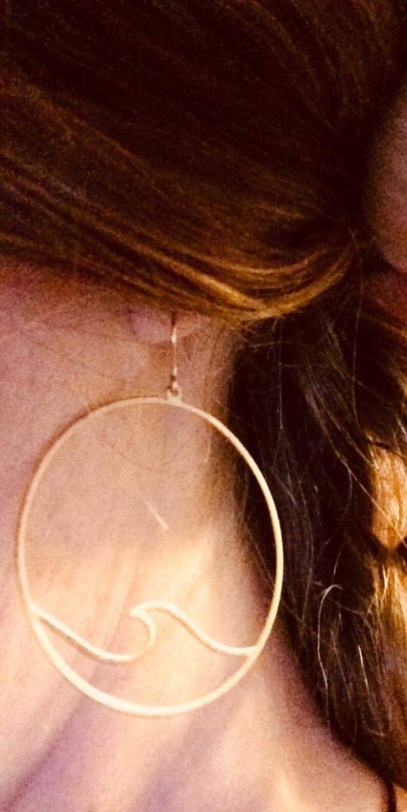 May include: Gold-colored hoop earrings with a wave design. The earrings are circular with a delicate wave detail cut out of the metal. The earrings are shown against a background of brown hair.