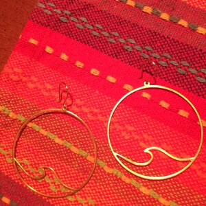 May include: Two gold-colored hoop earrings with a wave design. The earrings are displayed on a red and orange woven textile background. The earrings are circular with a wave design on the bottom.