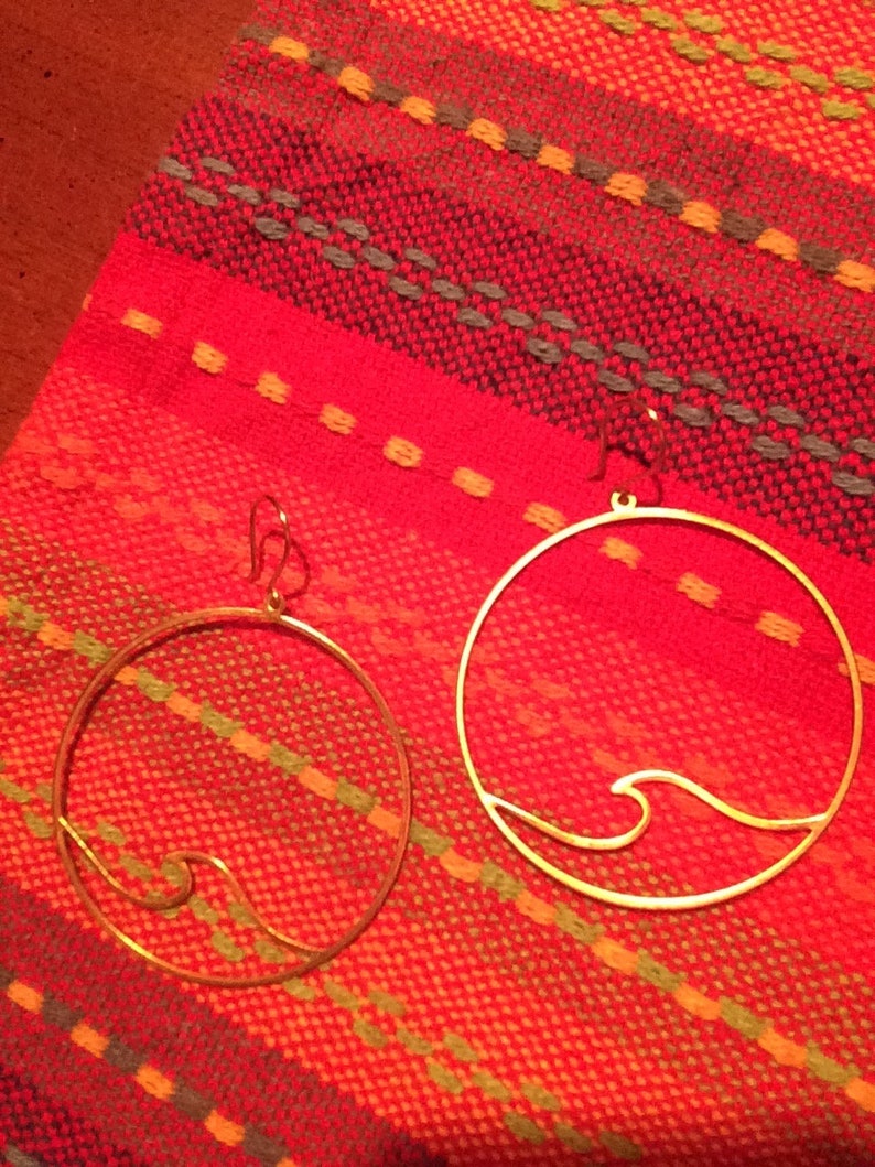 May include: Two gold-colored hoop earrings with a wave design. The earrings are displayed on a red and orange woven textile background. The earrings are circular with a wave design on the bottom.