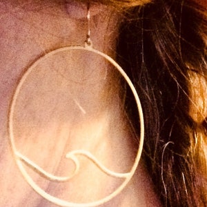 May include: A gold-colored, circular earring with a wave design. The earring is a simple, minimalist style, with a thin outline forming the circle and the wave. The earring is hanging from a small hook.