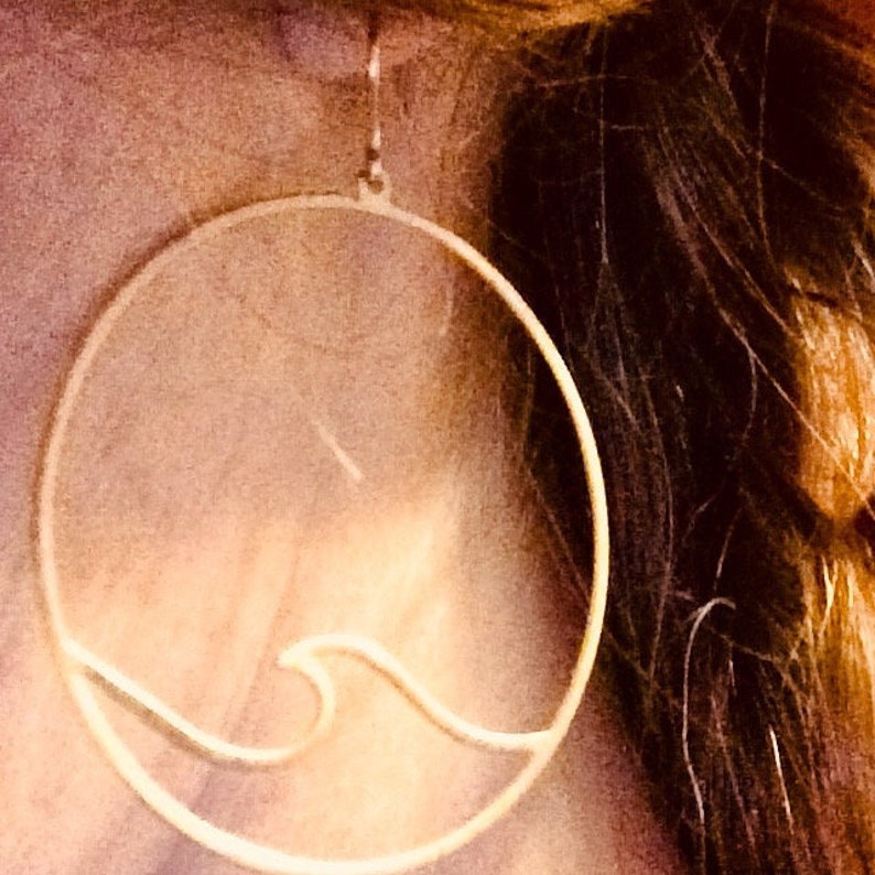 May include: A gold-colored, circular earring with a wave design. The earring is a simple, minimalist style, with a thin outline forming the circle and the wave. The earring is hanging from a small hook.