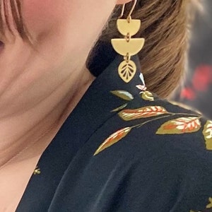 May include: Gold-coloured earrings with a geometric design. The earrings feature three stacked half-circle shapes and a leaf-shaped charm. The earrings are hanging from a gold hook. The person is wearing a black top with a floral pattern.