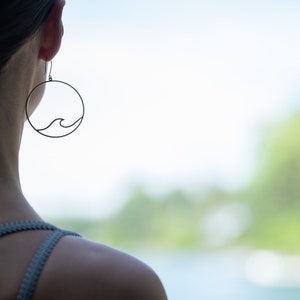 May include: A pair of delicate, black wave earrings. Each earring features a circular frame with a minimalist wave design. The earrings are shown worn on a person, with a blurred natural background.