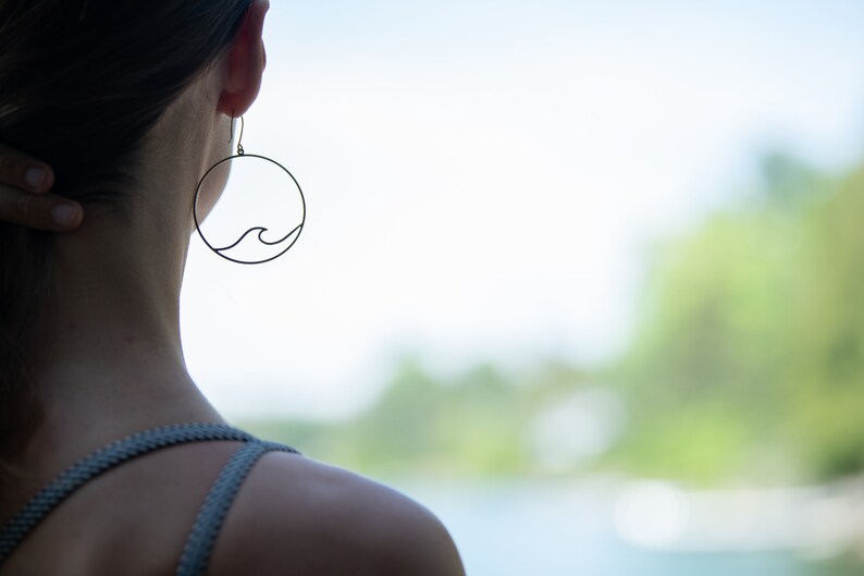 May include: A pair of delicate, black wave earrings. Each earring features a circular frame with a minimalist wave design. The earrings are shown worn on a person, with a blurred natural background.
