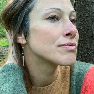 May include: A woman wearing a brown, green, and red sweater with a gold bar earring with green, red, and brown beads.