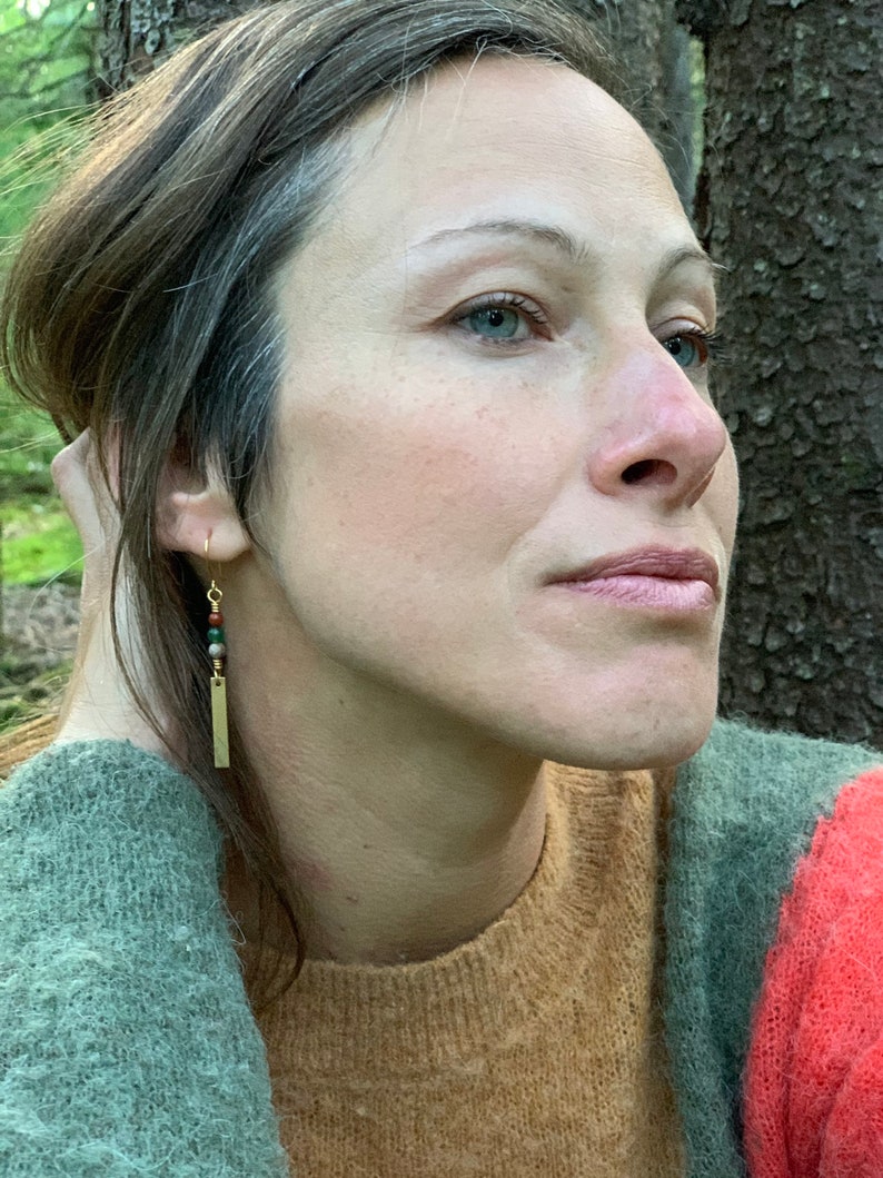 May include: A woman wearing a brown, green, and red sweater with a gold bar earring with green, red, and brown beads.