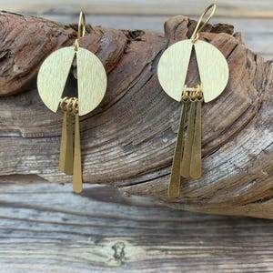 May include: A pair of gold-colored earrings. Each earring features a circular top with a triangular cutout and three dangling rectangular pendants. The earrings are displayed against a textured wooden background.