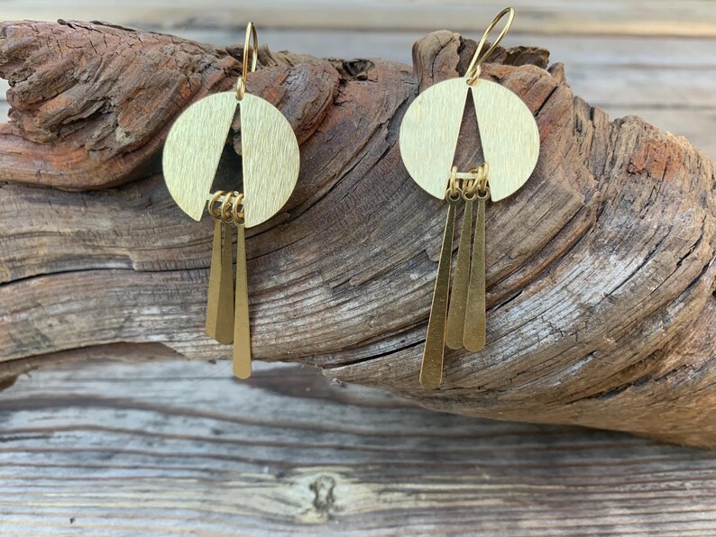 May include: A pair of gold-colored earrings. Each earring features a circular top with a triangular cutout and three dangling rectangular pendants. The earrings are displayed against a textured wooden background.