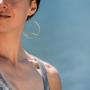May include: Gold-colored hoop earrings with a wave design. The earrings are circular, with a thin wave detail inside the circle. The background is a soft blue, suggesting a beach or water setting. The earrings are a jewelry item.
