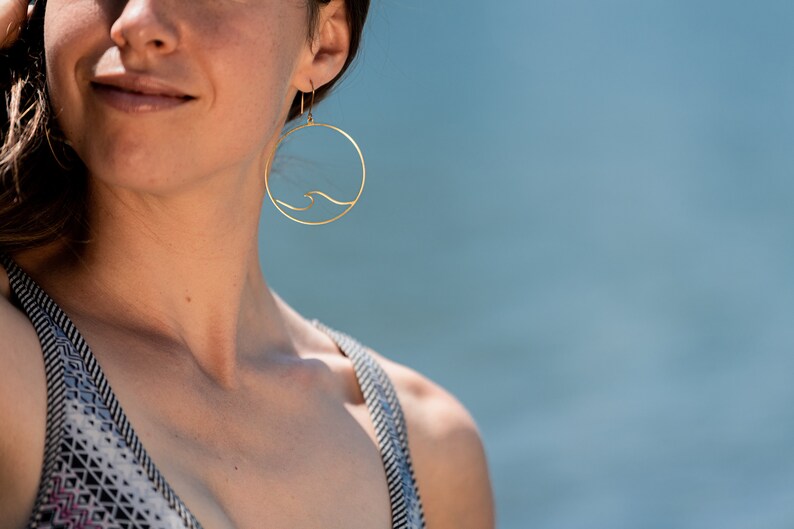 May include: Gold-colored hoop earrings with a wave design. The earrings are circular, with a thin wave detail inside the circle. The background is a soft blue, suggesting a beach or water setting. The earrings are a jewelry item.