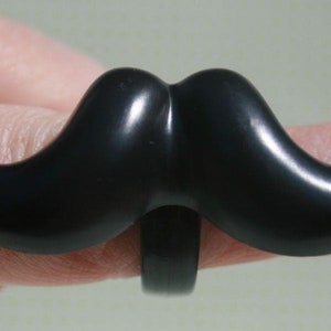 May include: A black mustache ring. The ring features a glossy, curved mustache design. The mustache is a bold, solid black color, and the ring band is also black. The ring is worn on a finger.