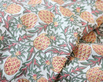May include: Fabric with a repeating pattern of yellow pineapples, green leaves, and pink accents on a light blue background. The design is a close-up shot of the fabric, showing the details of the print.