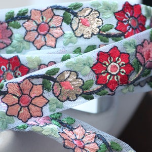 May include: White tulle trim with embroidered floral design in red, pink, and green. The flowers are arranged in a vine pattern.