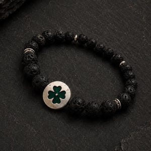 May include: A beaded bracelet featuring black porous lava stone beads and a circular silver-toned metal charm with a green four-leaf clover inlay. The design includes small silver-toned metal spacer beads interspersed among the dark textured stones.
