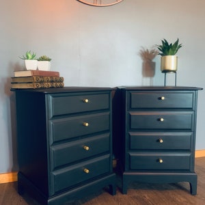 May include: Two black chest of drawers with gold knobs. Each chest has five drawers. The chests are on a wooden floor.