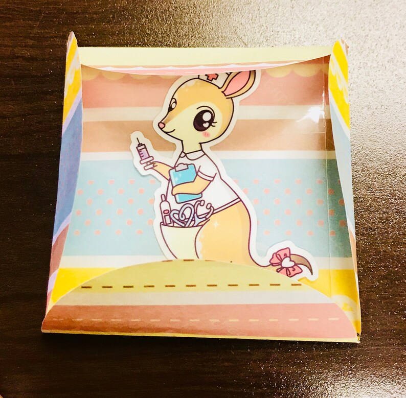 May include: A yellow and pink card featuring a cartoon kangaroo dressed as a nurse, holding a syringe. The kangaroo is surrounded by medical supplies, including a stethoscope and a bandage. The card has a white background with pink and blue stripes.