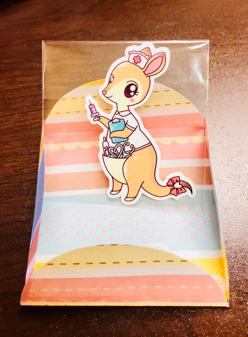 May include: A cute cartoon kangaroo wearing a nurse's uniform and a red cross hat. The kangaroo is holding a syringe and has a stethoscope and other medical supplies in its pouch. The image is on a colourful background with stripes and polka dots.