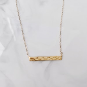 Op de afbeelding: Een gouden ketting met een staafvormige hanger en een delicate ketting. De staafvormige hanger heeft een gehamerde textuur.