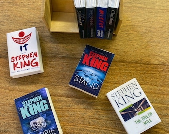 May include: Miniature Stephen King book collection featuring titles like "IT," "Carrie," and "The Stand." The books are arranged on a wooden surface, with some standing upright in a small wooden box. The covers display various colors and designs.
