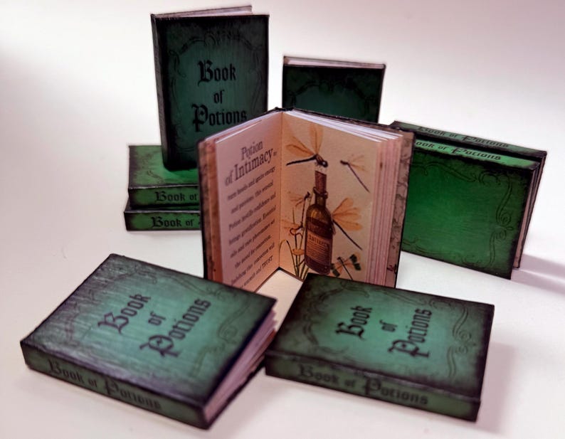Pu&ograve; includere: Mini libri con copertine verdi e il titolo "Book of Potions" in un carattere ornato. Un libro &egrave; aperto, rivelando illustrazioni di libellule e una bottiglia di pozione. I libri sono disposti su una superficie bianca.