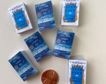 May include: Miniature books with blue and white covers are scattered across a surface. The titles "All the Light We Cannot See" and "Cloud Cuckoo Land" are visible. A penny is included for scale, showing the small size of the books.