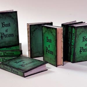 Pu&ograve; includere: Una collezione di libri in miniatura con copertine verdi e il titolo "Book of Potions" in caratteri gotici neri. I libri sono disposti in varie posizioni, alcuni impilati e altri in piedi.