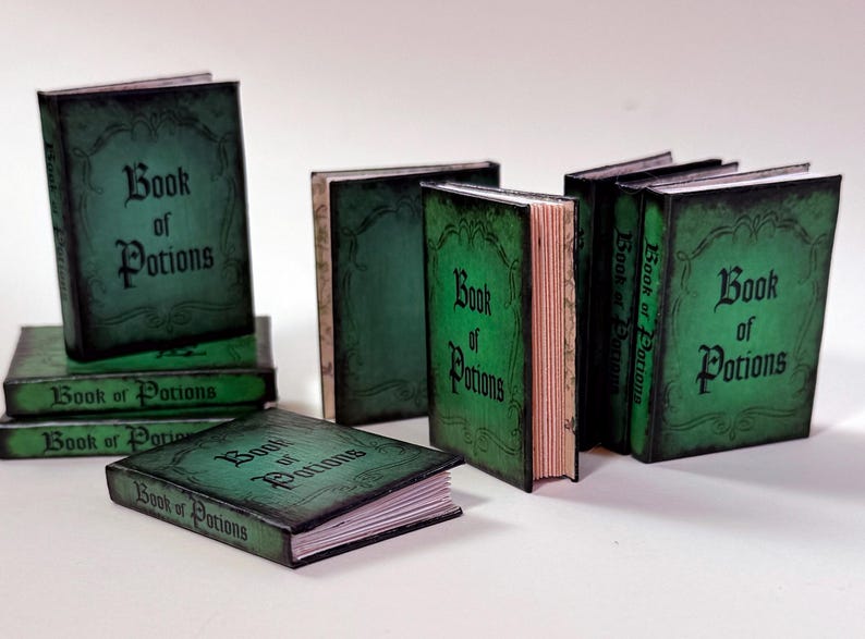 Pu&ograve; includere: Una collezione di libri in miniatura con copertine verdi e il titolo "Book of Potions" in caratteri gotici neri. I libri sono disposti in varie posizioni, alcuni impilati e altri in piedi.