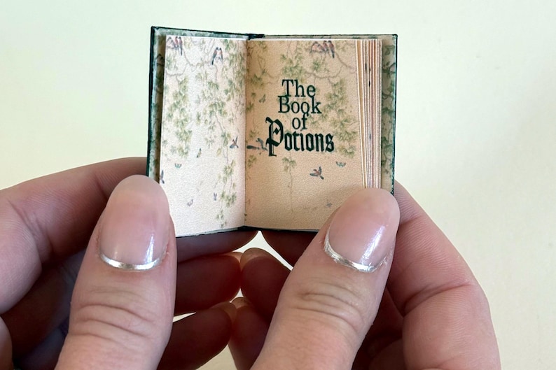 Pu&ograve; includere: Un libro in miniatura aperto intitolato "The Book of Potions". Le pagine hanno un'estetica vintage con illustrazioni botaniche. La copertina &egrave; verde scuro. Il libro &egrave; tenuto tra due mani.