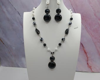 May include: A black and white beaded necklace and matching earrings set displayed on a white bust. The necklace features a mix of black, white, and patterned beads. The earrings are composed of two black circular beads.