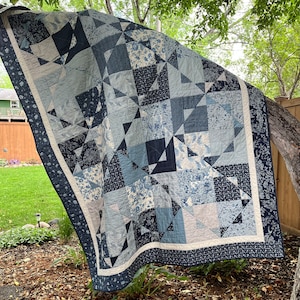 May include: A blue and white quilt with a geometric pattern. The quilt is made of various fabrics with floral and geometric designs. The quilt is hanging on a tree branch.
